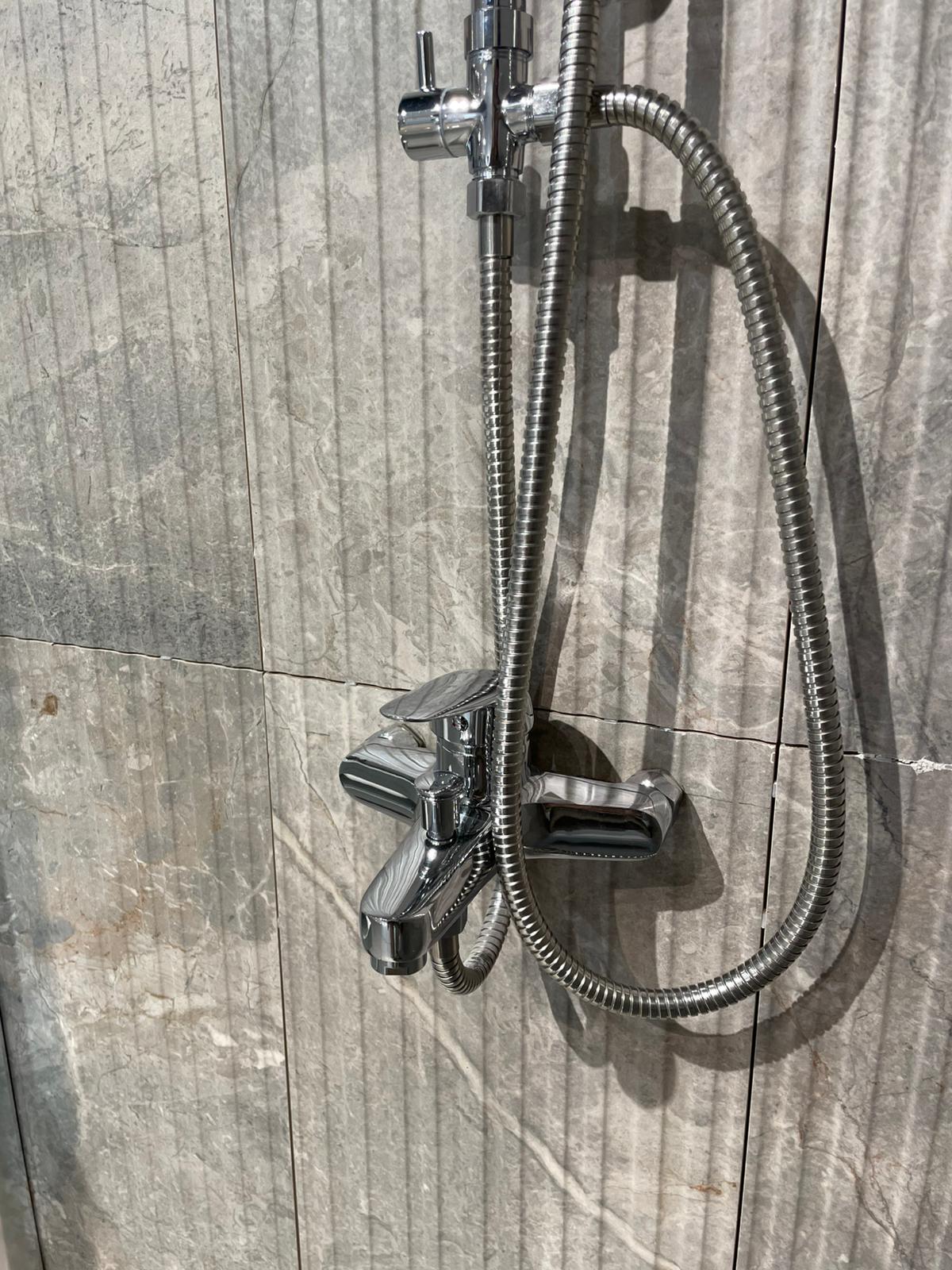 Moderna tuš baterija sa sjajnim hromiranim završetkom, postavljena na zid od prirodnog kamena. Ova oprema pruža funkcionalnost i estetski doprinos svakom kupatilu.